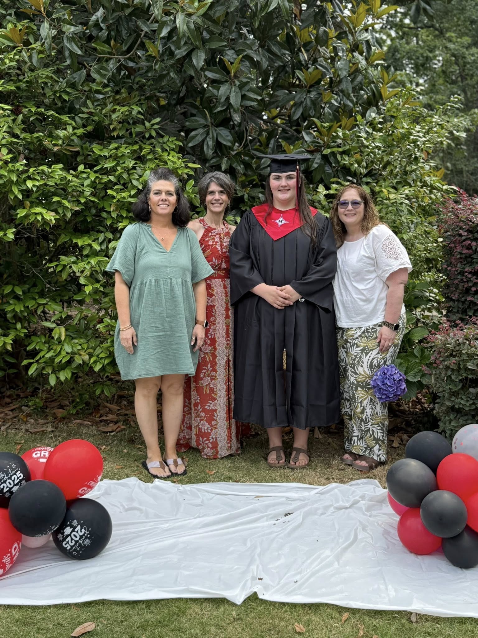 Four of us around the graduate, cap and gown, balloons on the grass.