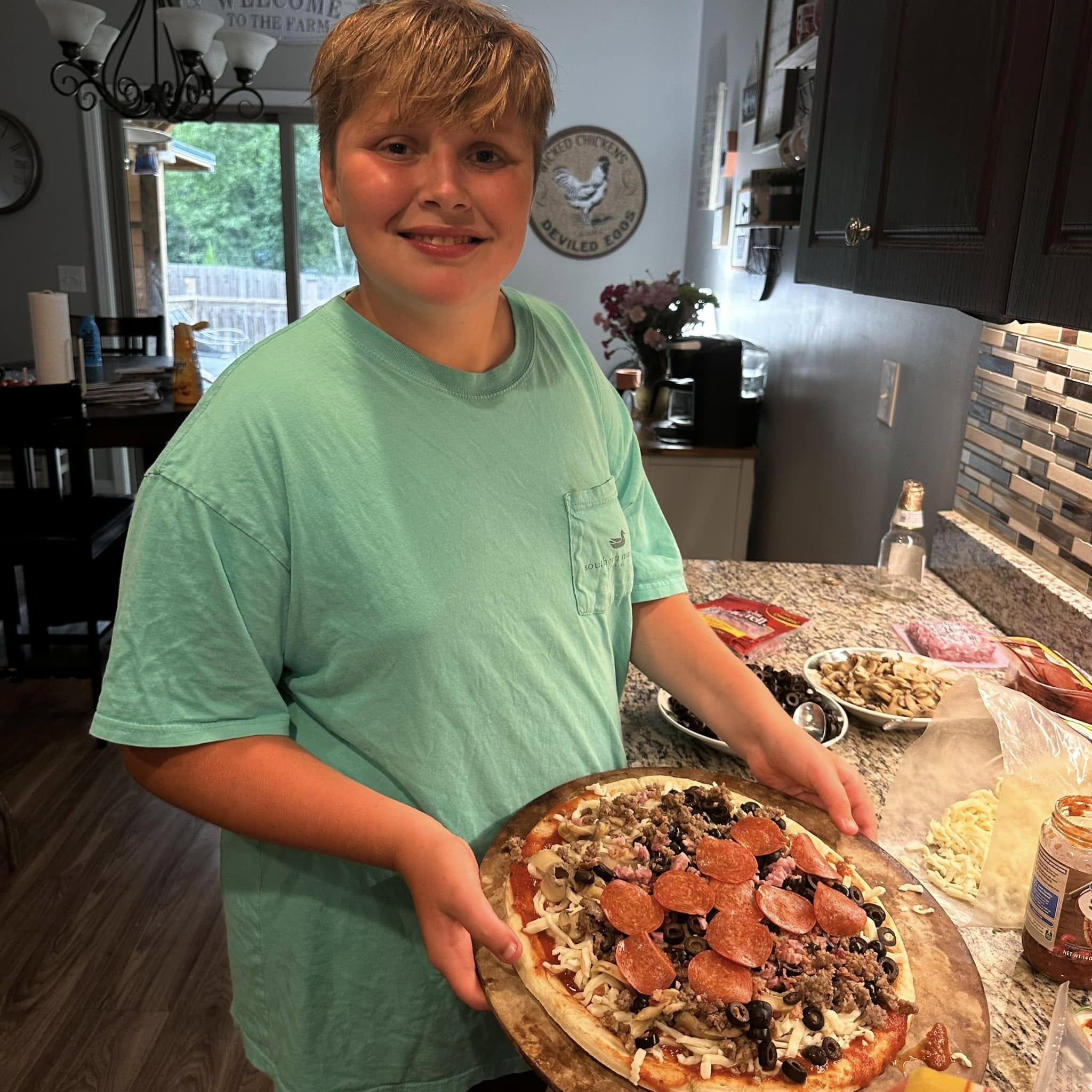 A teenager holding up a homemade pizza they built themselves in our kitchen.